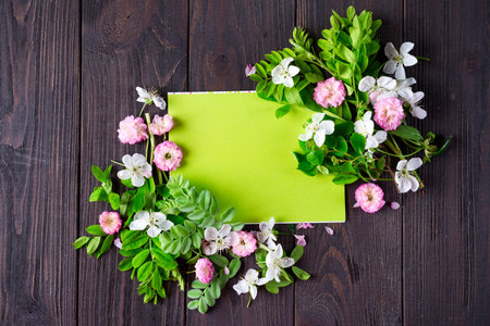 Frame of beautiful flowers with green paper copy space on brown wooden background, flat lay