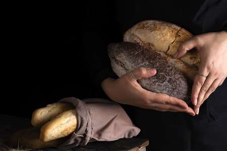 Close-up of woman hands as heart take fresh bread. Dark photo. Baking and cooking concept background. Hands of baker carefully hold loaf on wooden table, sprinkled with flour.の写真素材