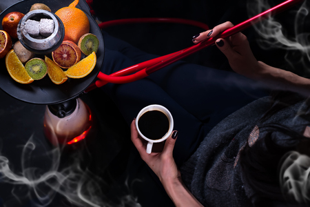 A girl with a cup of coffee in her hands smokes a hookah with fruit tobacco on a black backgroundの写真素材