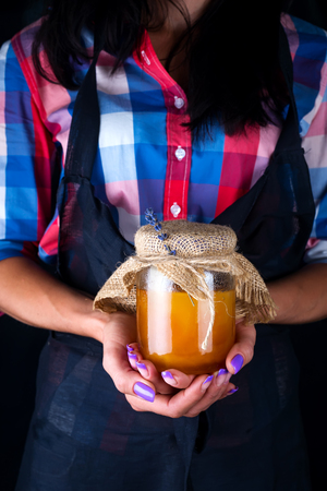 A fresh organic honey in a jar of a woman holds in hands on a dark background. Healthy foodの写真素材