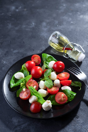 Delicious caprese salad with ripe cherry tomatoes and mini mozzarella cheese balls with fresh basil leaves on black plate and dark slate background. Italian healthy food conceptの写真素材