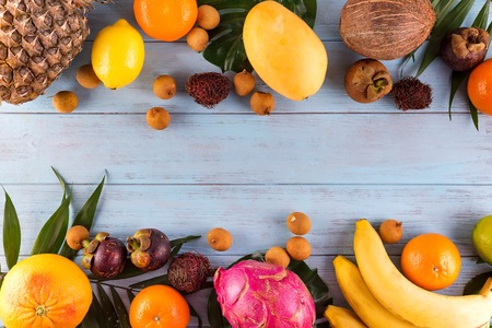 Healthy rainbow tropical fruits frame with palm leaves on blue wooden table, top view, copy space