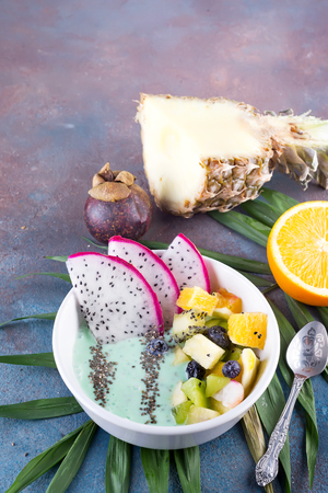 Breakfast kiwi smoothie bowl topped with pitaya, pineapple, chia seeds and berries with palm leaf on stoneの写真素材