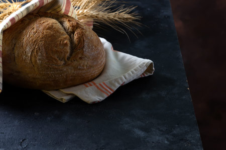 Round freshly baked rustic rye round bread with wheat ears and napkin on a dark background, copy...