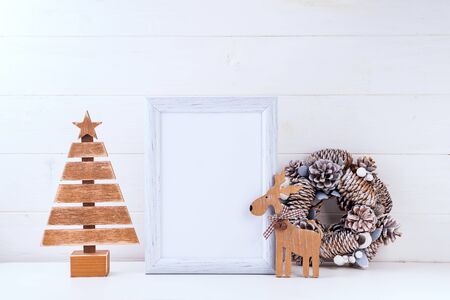 Christmas mockup with white frame, wreath of cones, wooden tree and deer on white wooden background. Christmas conceptの写真素材