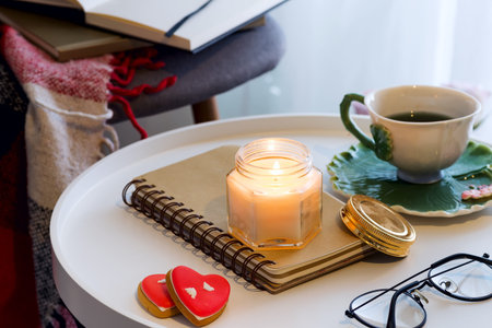 A candle with a notebook, cup of tea on a table with a chair, a plaid and a book. Home interior