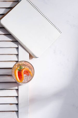 Cold summer homemade cocktail in a glass with slice of grapefruit and and paper note book on a white background with part of wooden jalousie. Top view.の写真素材