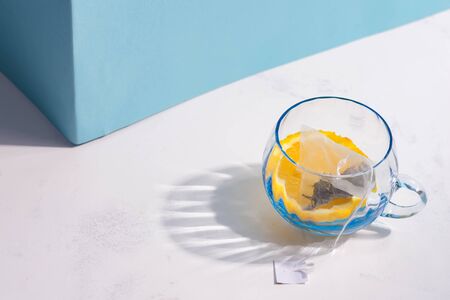 Glass cup with tea bag and slice of lemon for preparation cold natural summer homemade fruit tea drinks on a white table against blue wall with long shadow, copy space. Non-alcoholic summer drink.の写真素材