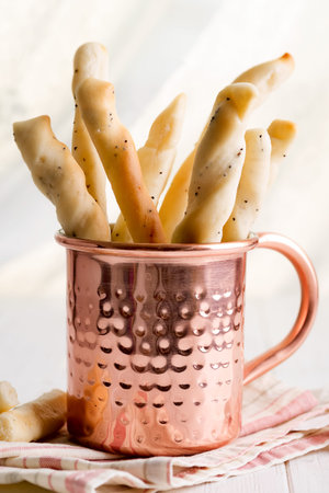 Traditional Italian bread sticks with poppy seeds in a copper cup on a textile napkin and wooden