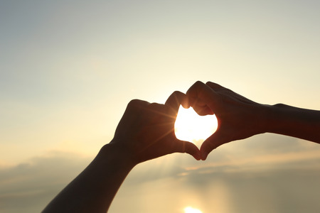 Heart shape making of hands against bright sea sunrise and sunny golden way at waterの写真素材