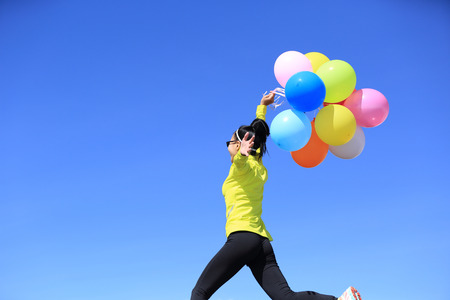 cheering young woman with colorful balloons jumping against blue skyの写真素材