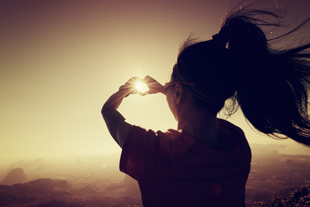 silhouette of successful woman hands making a heart shape on sunrise mountain topの写真素材
