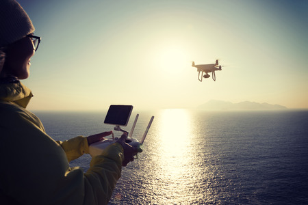 woman remote controlling a flying drone which taking photo over sunrise seaの写真素材