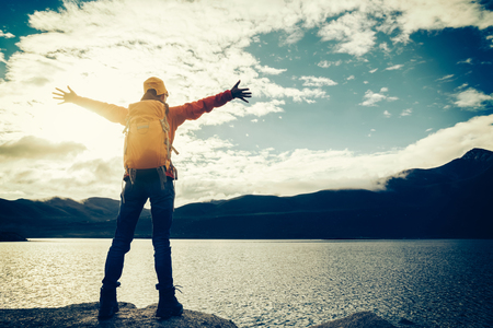 cheering young backpacking woman hiker outstretched arms face the mountains on watersideの写真素材