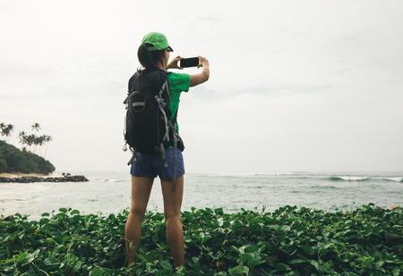 Young woman hiker using smartphone taking photo at seasideの写真素材