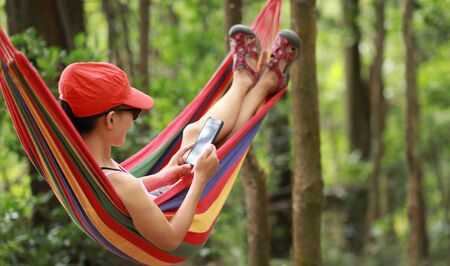 Woman hiker relaxing in hammock with smartphone in forestの写真素材