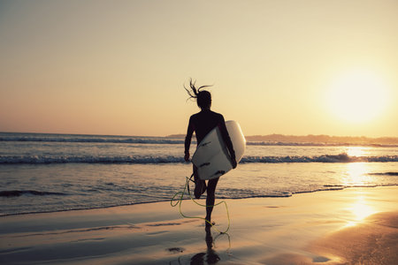 Female surfer walking with surfboard on the beach at sunsetの写真素材