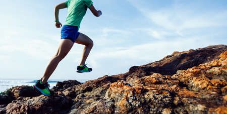 Woman trail runner running to rocky mountain top on sunrise seasideの写真素材