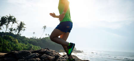 Woman trail runner running to rocky mountain top on sunrise seasideの写真素材