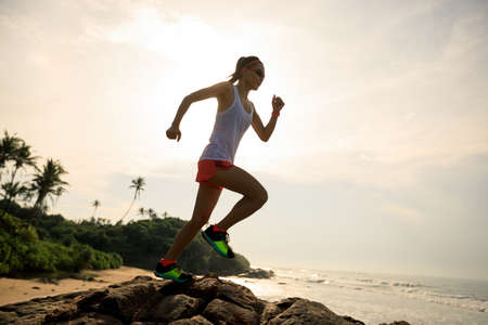 Woman trail runner running to rocky mountain top on sunrise seasideの写真素材