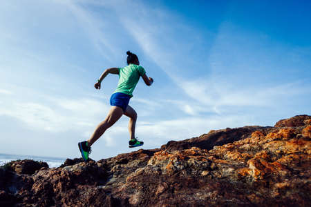 Woman trail runner running to rocky mountain top on sunrise seasideの写真素材