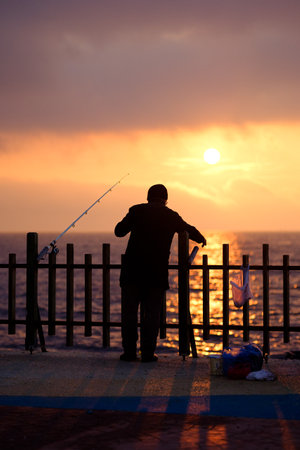 A fisherman leans on a fence, gazing at the sea as the golden sunset reflects on the water, casting long shadows. His fishing rod rests beside him, evoking solitude and tranquilityの写真素材