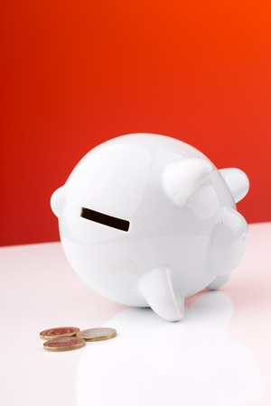 White piggy bank on a red background: it's on its side with a few penny's, a concept of a tight budget. The image is tilted.の写真素材