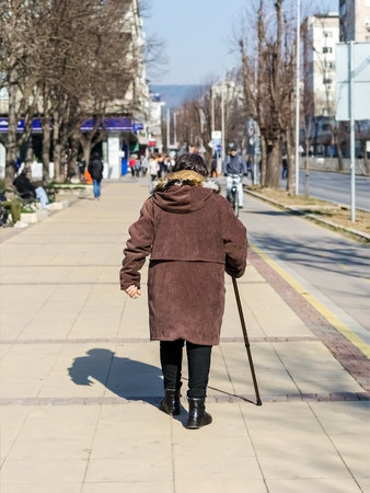 Foto de Varna, Bulgaria, February 20, 2019. An elderly woman dressed a brown jacket and with a cane in her hand walking along a city street on a sunny day, back view. - Imagen libre de derechos