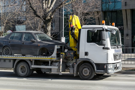 Varna, Bulgaria, February 28, 2019. A tow truck with hook and chain transports a car without a front wheel along the city street during the day. The concept of car evacuation. City life.のeditorial素材