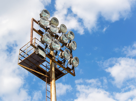 Twelve powerful spotlights on a lighting mast over the stadium against the blue sky with white...