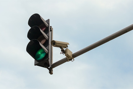 Outdoor surveillance camera and green traffic light both installed on a pole above a roadway, against a blue sky. Modern automatic traffic control. Copy space.