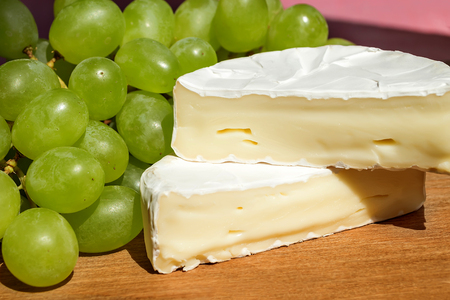 Pieces of tasty cheese camembert and sweet green grapes under a sunlight from the window on a brown wooden background. Soft cheese covered with edible white mold.の写真素材