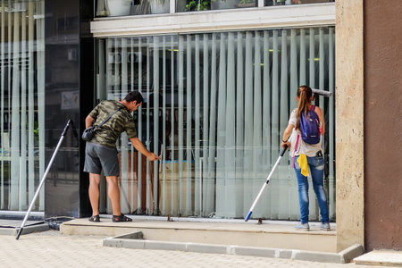 Varna, Bulgaria, June 11, 2019. Man and a woman with mops wash a large office window on the first floor of a building from the street side. Real people and city life.のeditorial素材