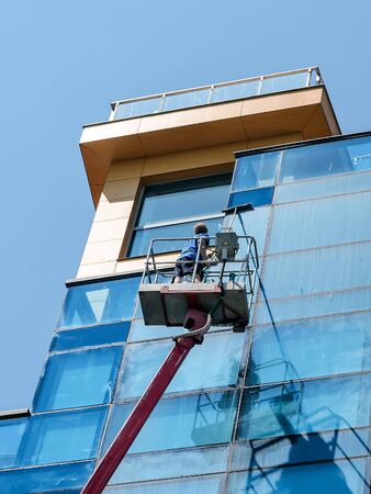 Man with mop washing a blue glass wall from autotower or aerial work platform on a sunny summer day. Real people and city life. Man washes a windows from autotower. Low angle view.の写真素材