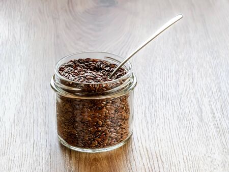 Raw flax seeds in a glass jar with metal spoon on a wooden table. Healthy food for lower cholesterol and preventing heart diseases and cancer. linseed is rich with antioxidants, vitamins, selenium and omega-3.  Front view.の写真素材