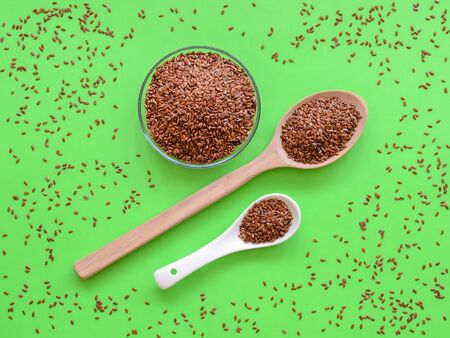 Wooden and ceramic spoons and glass bowl are full of whole flax seeds on a green background with scattered linseeds. Healthy food for lower cholesterol and preventing heart diseases. Top view.の写真素材