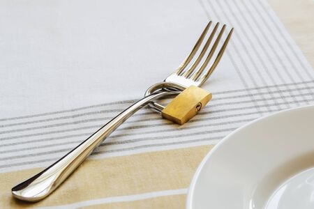Metal fork locked with padlock near white empty plate on a tablecloth. Control the amount of food eaten, losing weight and dieting consepts. Conscious food intake.の写真素材