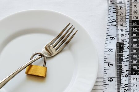 Metal fork locked with padlock on a white plate and measuring tape near it.  Control the amount of food eaten, losing weight and dieting consepts. Conscious food intake. Top view.の写真素材