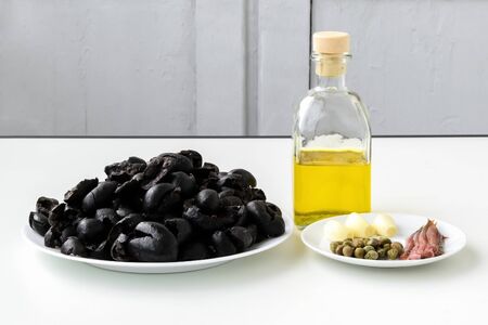 apenade ingredients: pitted black olives on white plate, olive oil in glass bottle, three cloves of garlic, some anchovy fillets and pickled capers on a sauser. Healthy eating. Front view.の写真素材
