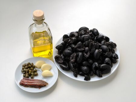 Ingredients for cooking tapenade: pitted black olives on white plate, olive oil in glass bottle, three cloves of garlic, some anchovy fillets and pickled capers on a sauser. Healthy eating. Top view.の写真素材