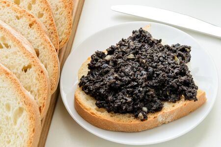 Tapenade sandwich on a sauser and sliced wheat bread on a wooden chopping board over white kitchen table. Homemade black olive paste with anchovies, garlic and olive oil. Top view.の写真素材