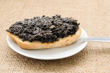 Fresh tapenade sandwich with slice of wheat bread in a white sauser and table knife on a burlap table napkin. Homemade black olive paste with anchovies, garlic and olive oil. Front view.の写真素材