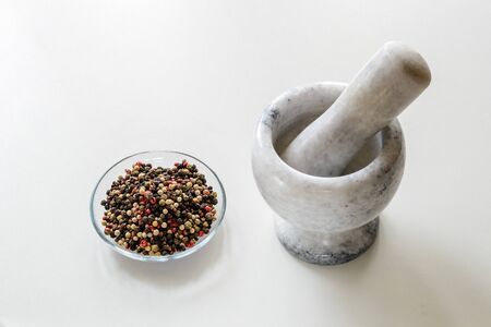 Glass saucer with mix pepper grains and marble or granite mortar with pestle for manual grinding spices on a white kitchen table. Modern utensil made of natural stone. Front view.の写真素材