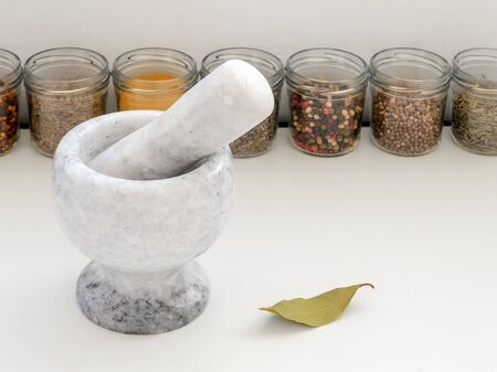 Marble or granite mortar with pestle for manual grinding and glass jars with different spices on a white kitchen table. Modern utensil made of natural stone. Front view.の写真素材