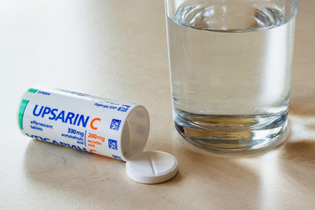 Varna, Bulgaria, October 02, 2019. Upsarin C antipyretic tablets and glass of water on a white table. Soluble effervescent aspirin with vitamin C pills for flu and cold treatment.のeditorial素材