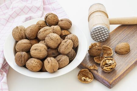 Natural unbleached walnuts (Juglans regia) in a white bowl. Cracked nuts and wooden meat mallet on a brown wooden board. Vegetarian food.の写真素材