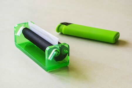 Green rolling machine with alwost done cigarette and green lighter on a white table. Making...