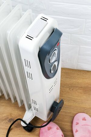 Pair of pink home slippers near oil-filled radiator in a room. White electric oil heater on a brown wooden floor. Home heating and comfort at winter. Top view.の写真素材