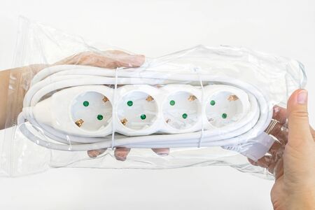 Woman hand holding new four outlets power extension cord in a transparent plastic pakage over white table. Electric power strip for home or office. Household appliances. Top view.の写真素材