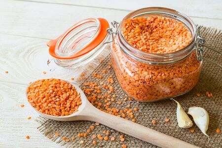 Dried red lentils in a glass clip top jar and wooden spoon on sackcloth table napkin over white...